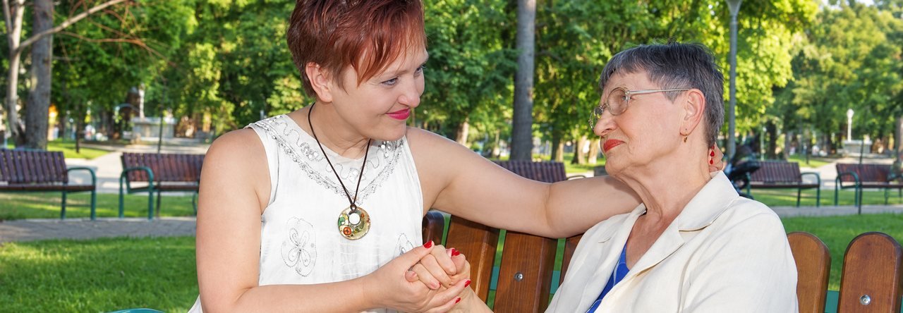 Социален асистент държи ръката на възрастна жена и разговарят на пейка в парка